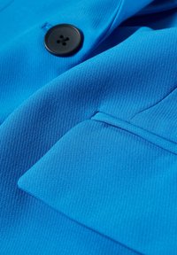 Tissu bleu à finition texturée, présentant un bouton noir proéminent et un détail de poche plaquée. Le matériau semble lisse et robuste.