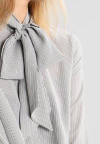 Randig blus i lätt tyg, med en stor rosett vid kragen och tunna vertikala ränder i växlande grå nyanser.