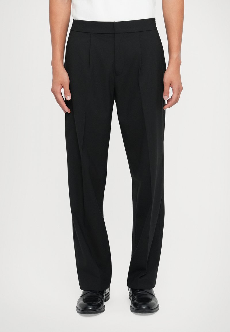 Black tailored trousers with a flat front, pleats, and a smooth texture, paired with polished black shoes; minimalistic design.