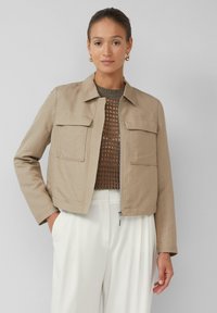 Vrouw in een beige ritsjack over een bruin mesh shirt en witte broek, staand met één hand in de zak tegen een effen grijze achtergrond.