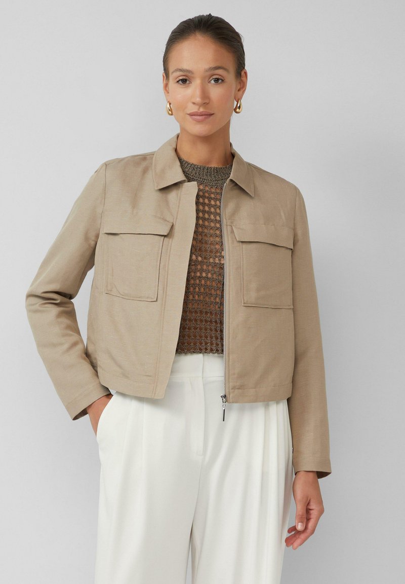 Vrouw in een beige ritsjack over een bruin mesh shirt en witte broek, staand met één hand in de zak tegen een effen grijze achtergrond.