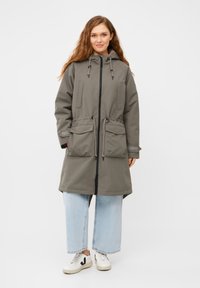 Impermeabile verde a metà lunghezza con cappuccio, vita con coulisse, due tasche frontali e cerniera nera. Indossato sopra jeans a gamba larga blu chiaro e sneakers.