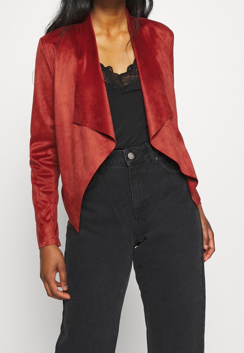 Femme portant une veste rouge à devant ouvert et drapé, un haut noir orné de dentelle et un jean noir taille haute, sur fond blanc.