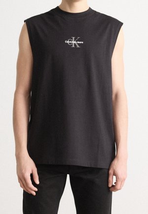 Osoba nosząca czarny top bez rękawów z białym logo "Calvin Klein Jeans" na piersi na jednolitym tle.