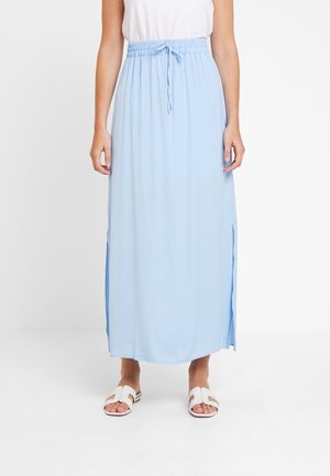 Femme portant une longue jupe bleu clair avec une taille élastique et des fentes sur les côtés, associée à des sandales blanches ouvertes et un haut blanc.
