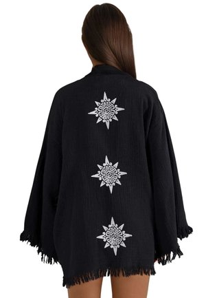 Frau mit langen braunen Haaren, die eine lockere schwarze Jacke trägt, die auf dem Rücken drei weiße, sonnenähnliche Muster und Fransen am Rand aufweist.