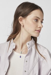 Collares de plata con piedras preciosas verdes y moradas cuelgan sobre una blusa color crema. Una camisa de lavanda clara tiene mangas abullonadas y botones a presión.