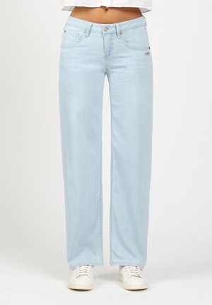 Woman wearing light blue wide-leg jeans and white sneakers, standing against a plain white background.