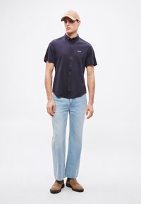 Tumesinise lühikeste varrukatega nööbitav särk, helesinised sirge lõikega teksad ja beež nokamüts logoga. Pruunad seemisnahast pahkluu saapad.