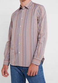 Mand iført en langærmet skjorte med knapper i smalle lodrette striber i nuancer af lilla, pink, beige og blå, parret med blå jeans.