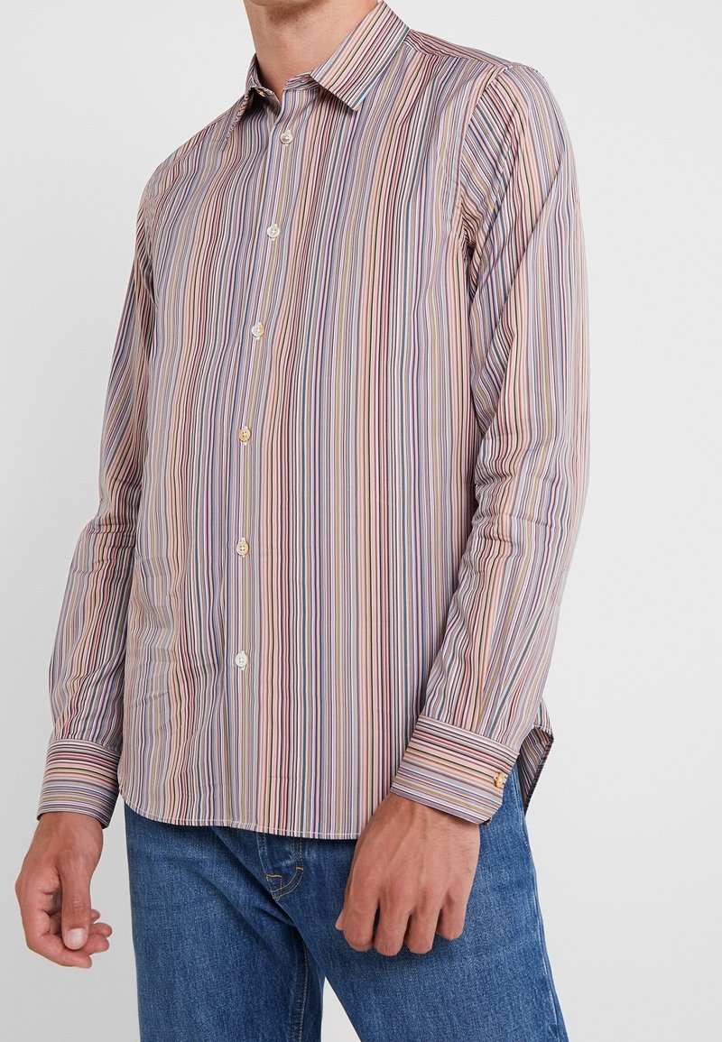 Mand iført en langærmet skjorte med knapper i smalle lodrette striber i nuancer af lilla, pink, beige og blå, parret med blå jeans.