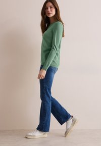 Pull vert à manches longues, manches cintrées, associé à un jean bleu taille mi-haute et des baskets blanches. Texture lisse, design minimaliste, coupe décontractée.
