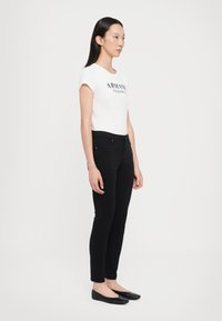 Melni šaurie džinsi apvienoti ar baltu pieguļošu T-kreklu, uz kura ir "Armani Exchange" zīmols. Valkāti ar melniem plakanajiem apaviem, uz balta fona.
