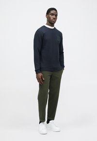 Marineblå sweatshirt med hvid krave, lavet af bomulds-materiale, med subtilt logo. Olivengrønne bukser i afslappet pasform og hvide sneakers.