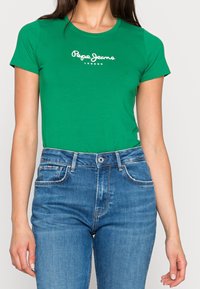 Grünes Baumwoll-T-Shirt mit kurzen Ärmeln, auf dem in weißem Schriftzug "Pepe Jeans London" steht, kombiniert mit blauen Jeans.