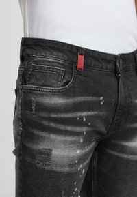 Czarne dżinsy z przetarciami, jasnym wyblaknięciem i smugami farby. Posiadają czerwoną metkę na pasku.