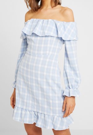 Vrouw draagt een lichtblauw-witte geruite off-shoulder jurk met lange opbollende mouwen en een gerimpelde zoom.