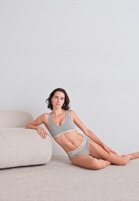 Moteris pilku Calvin Klein apatinį trikotažą vilkinti, ilsisi ant šviesios kiliminės dangos, remiasi į apvalų smėlio spalvos sofą prieš paprastą sieną.