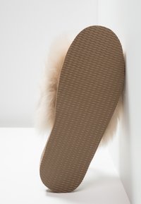 Chausson beige moelleux avec une semelle en caoutchouc texturée. La partie supérieure est douce et pelucheuse, avec un accent duveteux autour de la cheville.