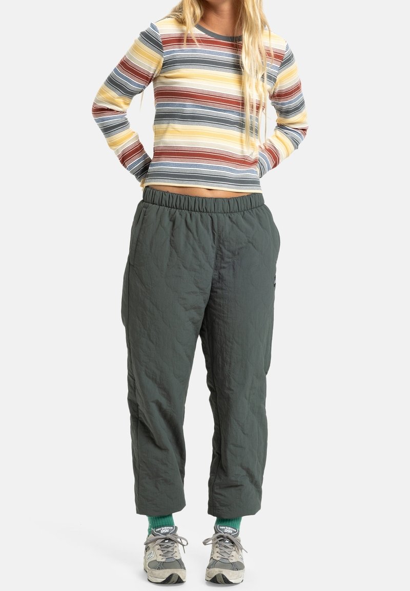 Femme portant un t-shirt à manches longues rayé multicolore, un pantalon ample vert foncé, des chaussettes vertes et des baskets grises, debout les mains dans le dos.