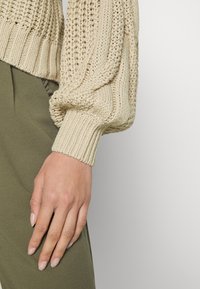 Beżowy sweter dziany z teksturowanymi rękawami i ściągaczami, w połączeniu z oliwkowymi spodniami, ukazujący zbliżenie na rękę i detale materiału.