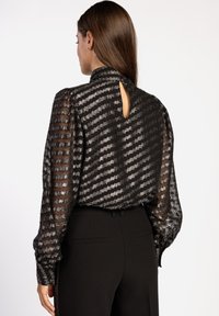 Zwarte, doorschijnende blouse met gestructureerde strepen, hoge kraag en sleuteltje aan de achterkant. Lange mouwen met elastische manchetten. Draagt samen met zwarte broeken.