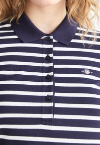 Marineblau- und weiße gestreifte Poloshirt aus gestricktem Stoff. Es verfügt über einen Kragen, sechs Knöpfe und ein kleines gesticktes Logo.