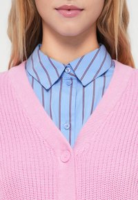 Cardigã rosa tricotado com decote em V e um botão rosa, sobre uma camisa azul clara com listras verticais estreitas e botões azuis.