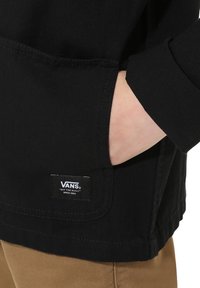 Czarna kurtka z kieszenią z przodu, prezentująca tkany wzór i stębnowane detale. Na kieszeni widoczna jest metka z logo Vans.