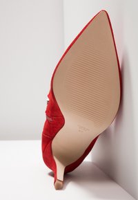 Chaussure à talon haut en daim rouge à bout pointu inclinée vers le haut, montrant une semelle beige texturée et un petit talon aiguilles sur fond blanc.