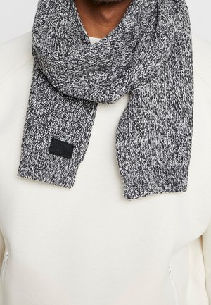 Écharpe en tricot gris et blanc enroulée autour du cou par-dessus un sweat-shirt blanc avec un petit logo rectangulaire noir sur l'extrémité de l'écharpe.