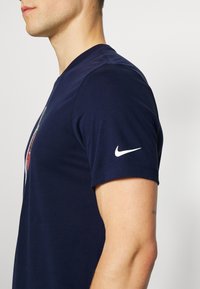 Námorně modré bavlněné tričko má kulatý výstřih, krátké rukávy a bílé logo Nike na rukávu; hladká textura a přiléhavý střih.