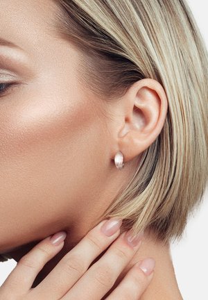 Ohrringe aus Roségold mit einem strukturierten, geschwungenen Design, getragen am Ohr eines Models, ergänzt durch helles Haar und natürliche Nägel.