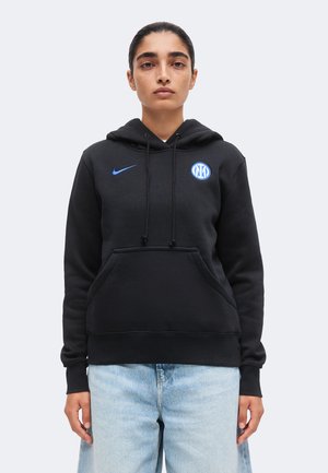 Személy fekete Nike kapucnis pulóvert visel kék Inter Milan logóval és világoskék farmernadrágot, egyszerű világos háttér előtt áll.