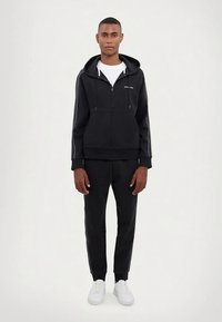 Male model wearing a black zip-up hoodie and matching black joggers with white sneakers, standing against a plain light background.