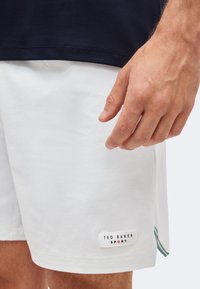 Κοντινό ενός χεριού κοντά σε λευκά αθλητικά σορτς Ted Baker με πράσινη ριγέ λεπτομέρεια στο πλάι.