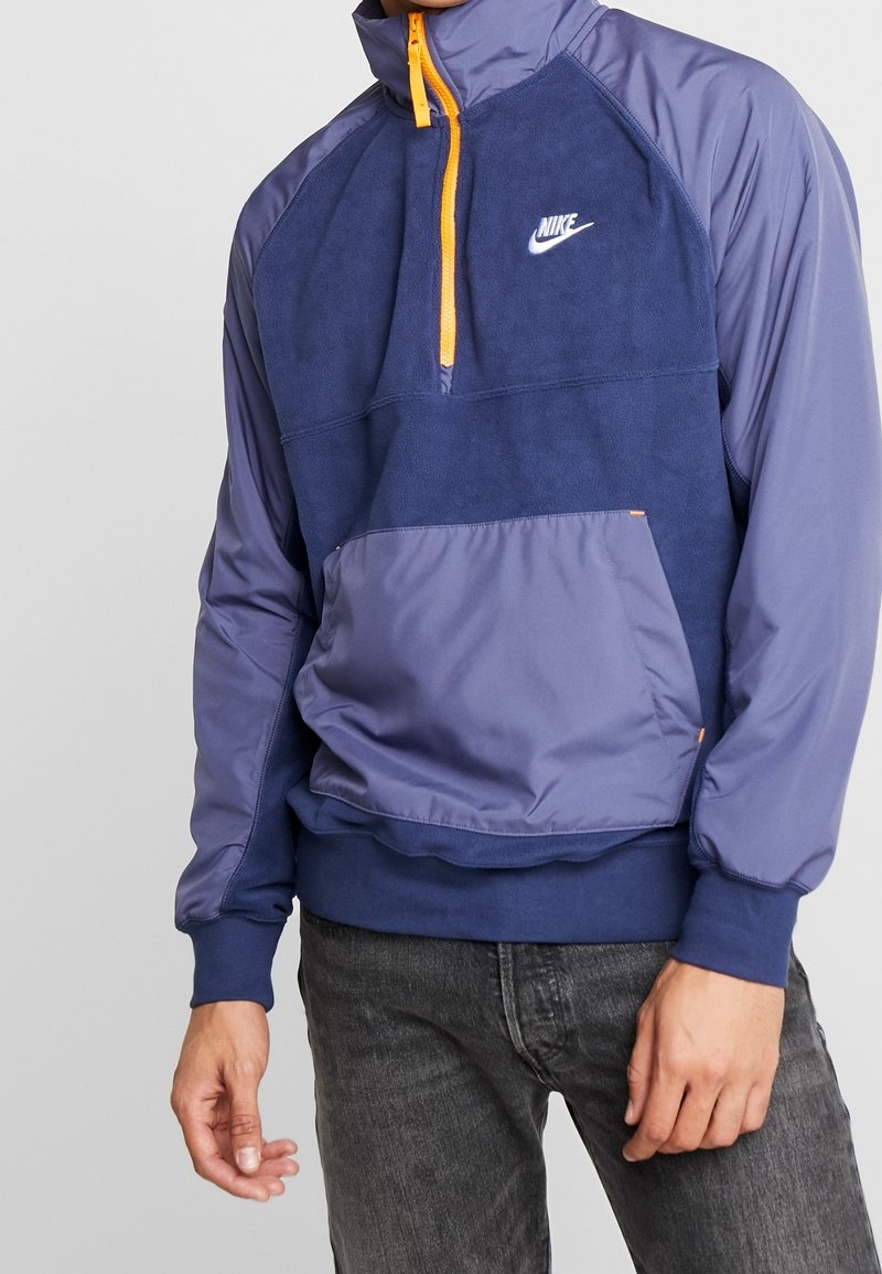 Nike halvlång dragkedjepullover i marinblått och lila. Har fleecematerial med elastisk fåll, frampocket och orange dragkedjedetaljer.