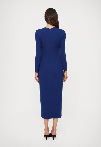 Mulher de costas, vestindo um vestido azul royal de mangas compridas e comprimento até ao tornozelo, com mules pretos de salto alto, contra um fundo branco.