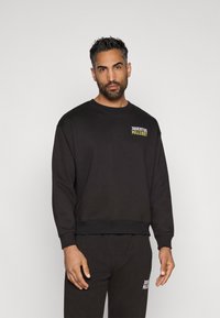 Sudadera negra de manga larga con puños acanalados, que presenta un pequeño logotipo multicolor de "JUVENTUS" en el pecho. Textura de tela suave.