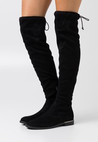Botas negras hasta la rodilla hechas de suave ante, con puntera redondeada, cremallera lateral y lazos decorativos en la parte superior, con suela plana.