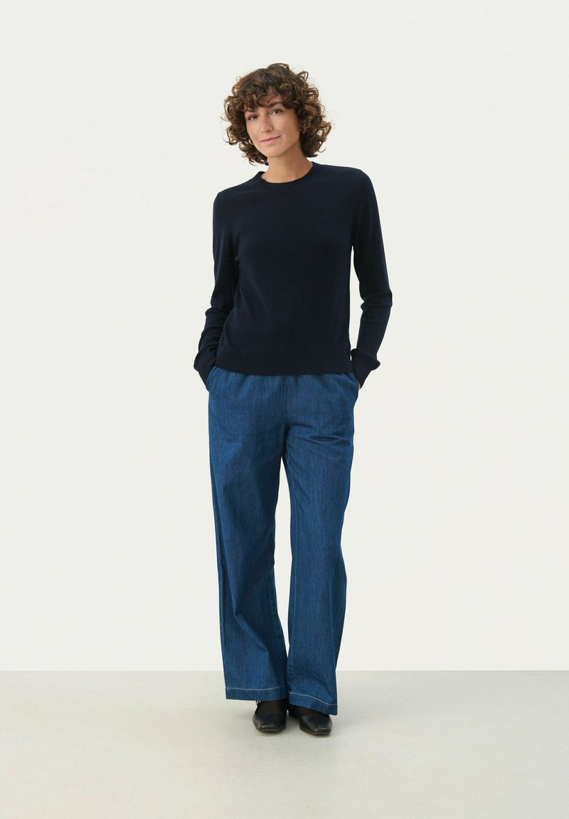 Marineblauer, langärmeliger Strickpullover kombiniert mit lockeren, hochgeschnittenen blauen Jeans. Glatte Textur mit seitlichen Taschen, abgerundet mit schwarzen Schuhen.