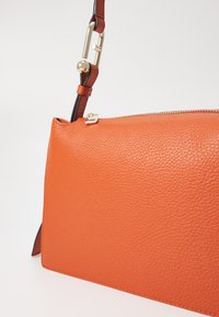 Orange läderhandväska med en texturerad yta, utrustad med en guldfärgad dragkedja och ett avtagbart brunt axelband.