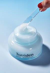 Valkoinen kermaisen pehmeä voide kirkkaassa, pyöreässä astiassa, jossa on vaaleansiniset kansi. Läpinäkyvä spatula nostaa kermaisesta voidetta.