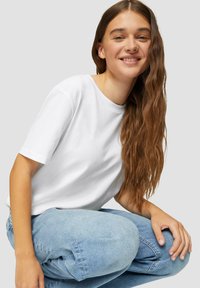 Witte katoenen t-shirt met korte mouwen, ronde halslijn en een ontspannen pasvorm, gecombineerd met lichtblauwe spijkerbroek. Soepele textuur, minimalistisch ontwerp.