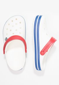 Tamancos brancos com tiras vermelhas, solas listradas em azul e branco, e orifícios de ventilação. Feitos de material em borracha com acabamento texturizado.