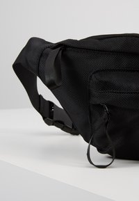 Bolsa de cintura de nylon negro con superficie texturizada, correa ajustable, bolsillos laterales con cremallera y cierre de hebilla seguro.