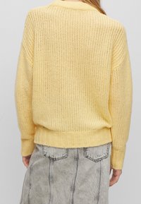 Stickad gul tröja med lös passform, ribbade muddar och kant, med ett texturerat vävmönster. Kombinerad med en grå jeanskjol som visar synliga sömmar.