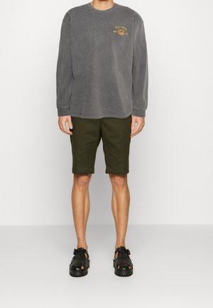 Maglietta grigia a maniche lunghe con logo ricamato, pantaloni corti verde oliva e sandali neri aperti con un design testurizzato.