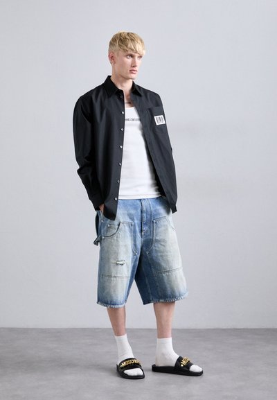 Ung mand iført en sort skjorte med knapper over en hvid tanktop, lyseblå baggy denimshorts, hvide sokker og sorte slides med guldtekst.