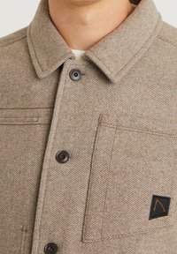 Brun strukturerad skjortjacka med spridd krage, som har bruna knappar och en svart lappar med en orange grafik.